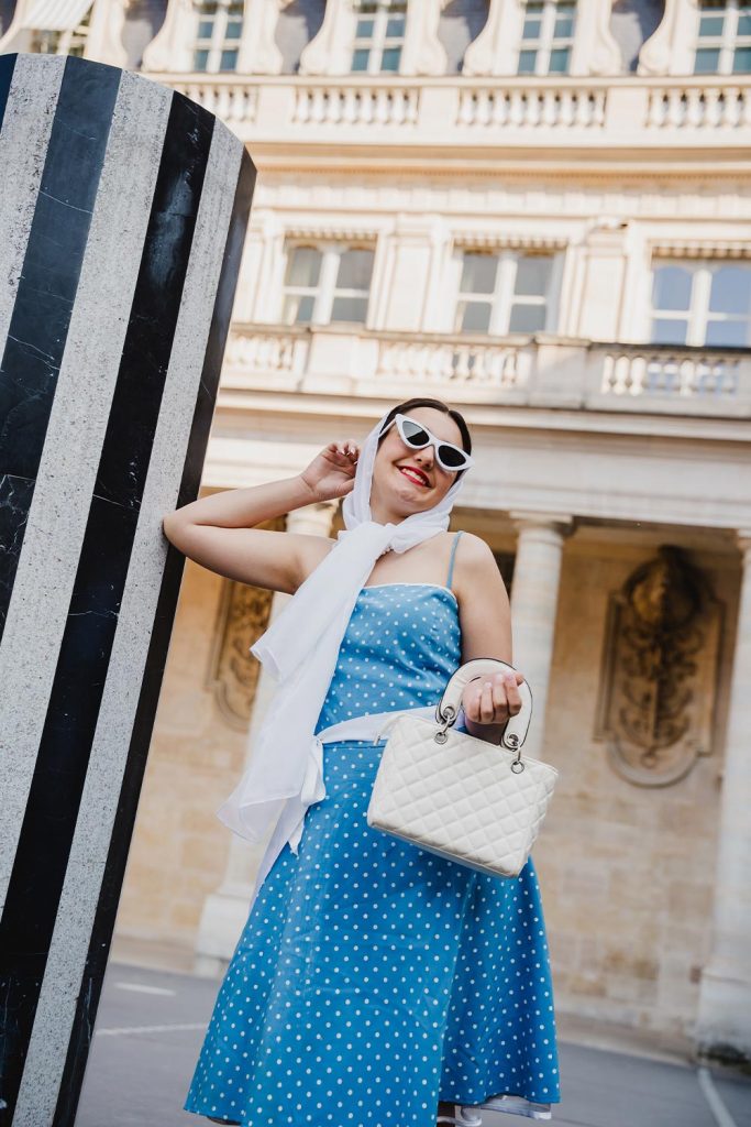 Quinceañera con vestido azul de lunares, pañuelo y gafas de sol estilo vintage en las columnas del Palais Royal, París / Quinceañera in a blue polka dot dress, headscarf, and vintage-style sunglasses at the Palais Royal columns, Paris
