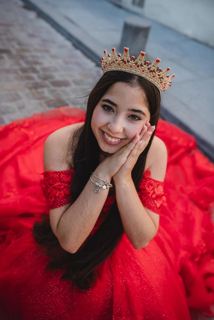 Quinceañera sonriendo desde arriba con vestido rojo voluminoso y corona de piedras rojas en una calle de París / Quinceañera smiling from above in a voluminous red gown and red-stone crown on a Paris street