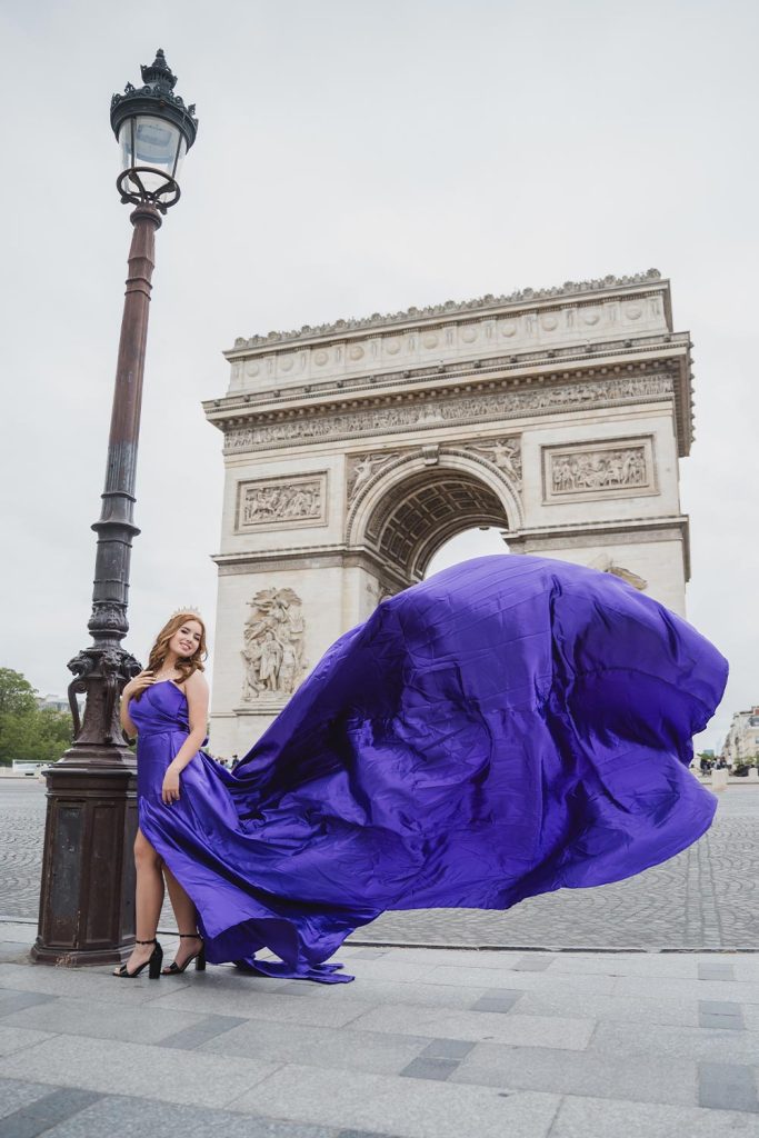 Quinceañera junto a un farol parisino con vestido morado de tela voladora frente al Arco de Triunfo / Quinceañera by a Parisian lamppost with a flowing purple gown in front of the Arc de Triomphe