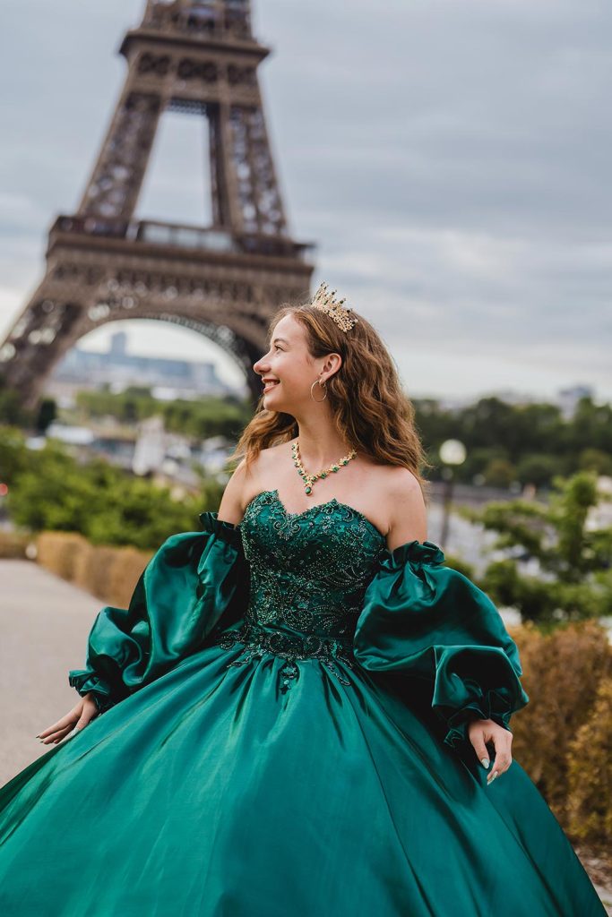 Quinceañera con vestido verde esmeralda de mangas abullonadas y corona dorada frente a la Torre Eiffel / Quinceañera in an emerald green gown with puffy sleeves and a gold crown in front of the Eiffel Tower