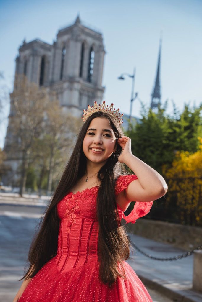 Quinceañera con vestido rojo bordado y corona sonriendo frente a la catedral de Notre Dame en París / Quinceañera in an embroidered red gown and crown smiling in front of Notre Dame cathedral in Paris