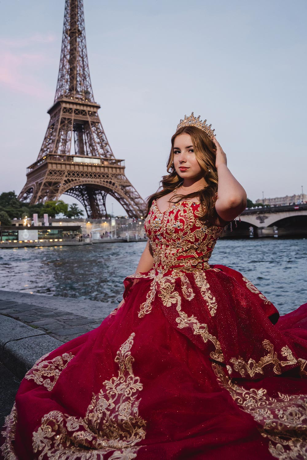 Quinceañera con vestido rojo y dorado bordado sentada junto al río Sena al atardecer con la Torre Eiffel de fondo / Quinceañera in an embroidered red and gold gown seated by the Seine at sunset with the Eiffel Tower behind